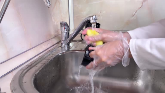 A person wearing gloves washing a kitchen faucet with a sponge, highlighting the ease of cleaning a fingerprint-resistant surface.